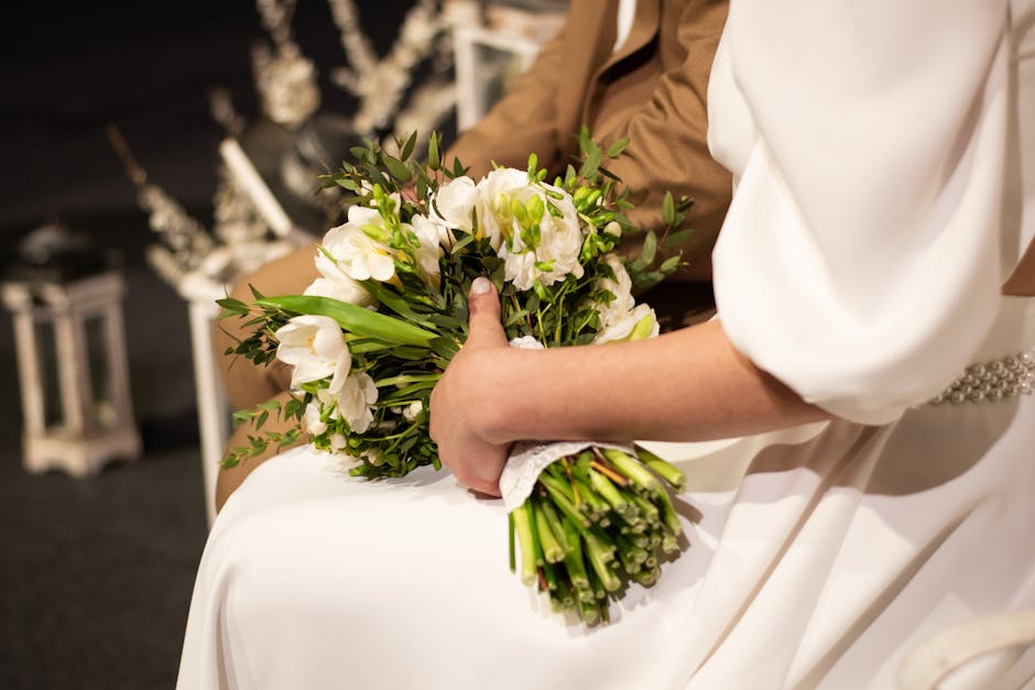 Close-up of a bride holding a large, round bouquet composed of soft pink and cream peonies and white roses, arranged with lush, ruffled petals and fresh green stems visible at the base. The bouquet is