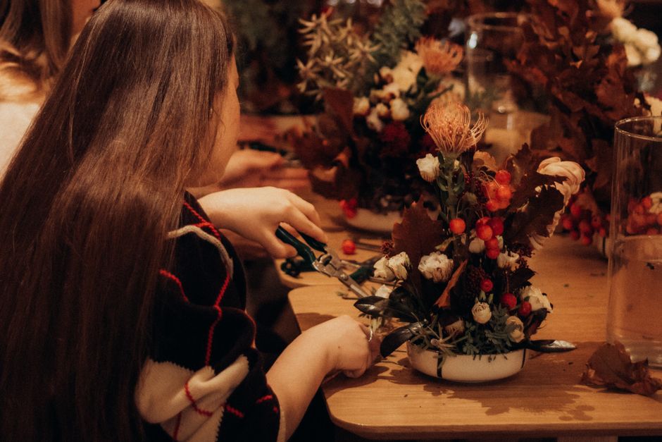 A florist's hand is seen arranging a floral centerpiece on a wooden table, featuring a variety of fresh flowers including pale cream roses, pink and peach-hued dahlias, and orange berries, accented wi