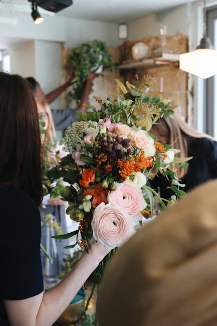 Close-up of hands preparing a floral arrangement with fresh flowers including pink and white tulips, and yellow roses, arranged on a dark surface. The person is holding a roll of natural twine in one 