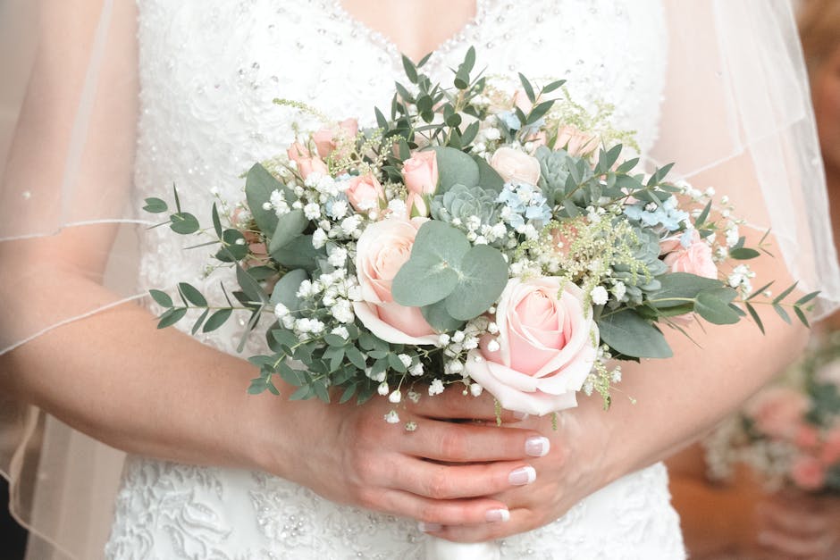 A person in a white sleeveless dress holds a large floral bouquet featuring cream and yellow flowers, including roses and daffodils, with green foliage accents. The bouquet is arranged in a loose, nat
