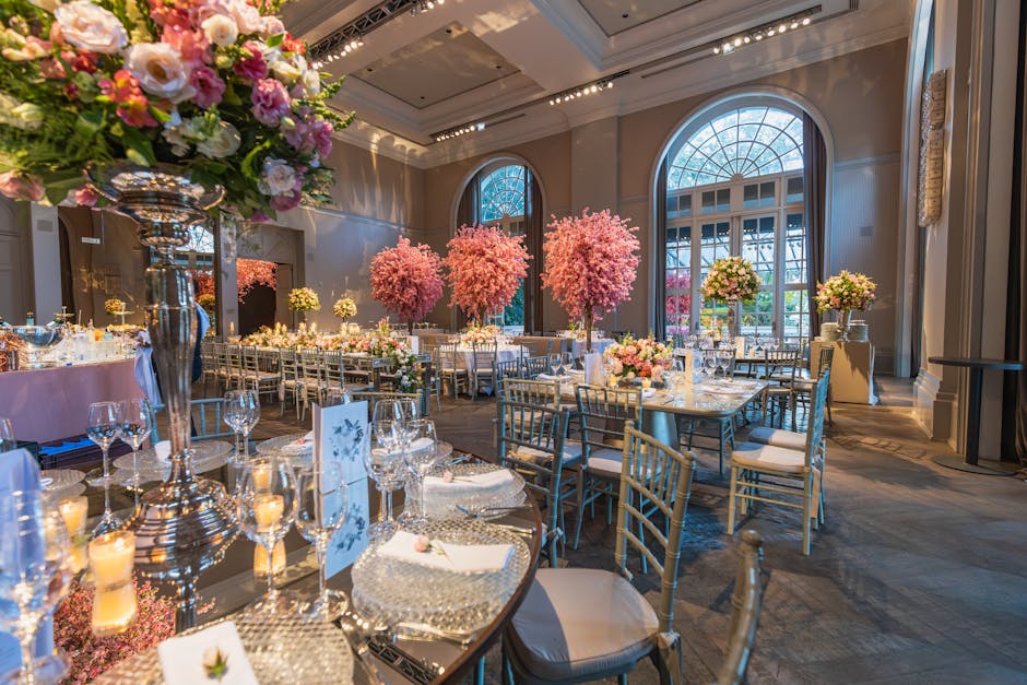 A luxurious event space featuring a large, vibrant pink cherry blossom tree with dense blooming branches situated on a circular pink velvet sofa, surrounded by smaller pink and white floral arrangemen